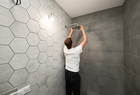 Back view of man standing by the wall with ceramic tile and installing high pressure bath showerhead at home. Male worker fixing bathroom showerhead while working on bathroom renovation.の写真素材
