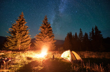 Night camping in mountains under starry sky and Milky way. Female tourist resting near burning campfire and illuminated tent in campsite, lying on grass and enjoying night sky with stars.の写真素材