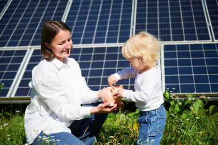Little child and his mother saving money and putting cash into piggy bank. Mom and her son putting savings into piggybank on background of solar panels. Concept of investment in renewable energy.の写真素材