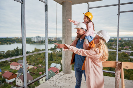 Man holding daughter and woman pointing at something while standing next to husband at construction site. Family with child future homeowners observing apartment building under construction.の写真素材