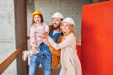 Man holding daughter and smiling while kid putting apartment keys in woman hand. Portrait of family with child standing in building under construction. Happy family life and new home purchase concept.の写真素材