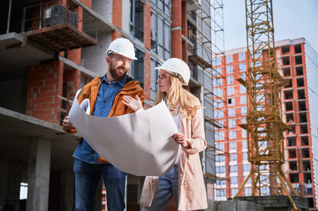 Couple in safety helmets studying architectural drawings outside apartment building under construction. Man and woman discussing building plan outdoors at construction site.の写真素材