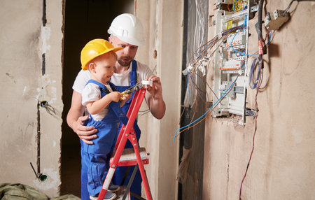 Toddler boy electrician standing in ladder and holding pliers tool while installing switchboard with father at home. Man and kid mounting electrical wiring in apartment under renovation.の写真素材