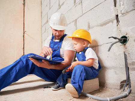 Male construction worker sitting by brick wall and pointing at clipboard while showing documents to child. Man and kid wearing safety helmets and work overalls while working on home renovation.の写真素材