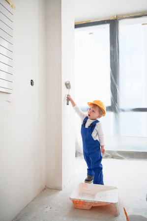 Child construction worker painting wall with paint brush in apartment. Kid in safety helmet and work overalls using paint brush while playing at home under renovation.の写真素材