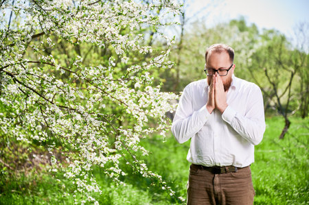 Man allergic suffering from seasonal allergy at spring in blossoming garden at springtime. Young bearded man with glasses in white shirt sneezing in front of blooming tree. Spring allergy conceptの写真素材