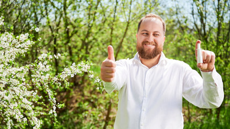 Man allergic using medical nasal drops, suffering from seasonal allergy at spring in blossoming garden. Portrait of smiling man showing thumbs up near blooming tree outdoors. Spring allergy concept.の写真素材