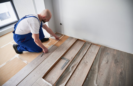 Man in work overalls using tape measure while installing laminate flooring in apartment under renovation. Male builder checking distance from wall to laminate board. Hardwood floor renovation concept.の写真素材