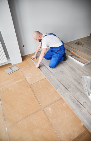 Man in workwear measuring distance from wall to laminate board with measuring tape ruler. Male construction worker using tape measure while installing laminate flooring in apartment under renovation.の写真素材