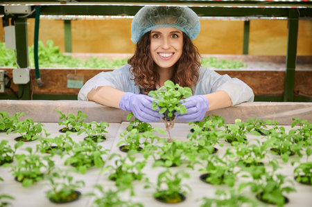 Female gardener working in greenhouse, holding pot with plant seedlings and smiling while standing near shelf with seed trays. Cheerful woman wearing disposable cap and garden gloves.の写真素材