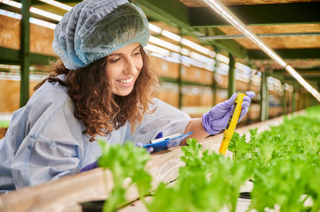 Woman scientist using pH meter in greenhouse. Joyful female gardener holding clipboard and smiling while measuring soil pH value with digital electronic device.の写真素材