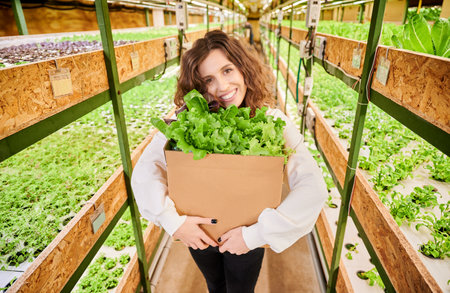 Happy female customer looking at camera and smiling while holding cardboard package box with green leafy plants. Woman with fresh leafy greens standing near shelves with plants in greenhouse.の写真素材