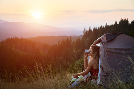 Woman hiker camping in mountains at sunset. Beautiful female sitting in tents entrance on grass with cup of tea. Young tourist hiking, having rest in campsite. Concept of tourism and hiking.の写真素材