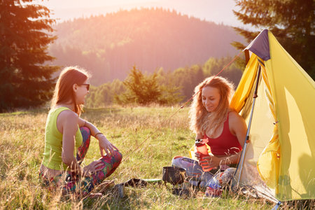 Two women tourists camping in mountains. Young women sitting in campsite, having break. Friends sitting on grass, talking, smiling, making tea. Concept of tourism and hiking.の写真素材
