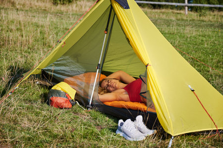 Woman tourist camping outdoors. Tired woman hiking in hills, lying in tent. Slim, blonde female having break, admiring landscape. Concept of freedom and beauty of nature.の写真素材
