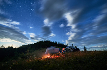Cozy camping in mountains under evening cloudy sky. Starry night above bonfire, tourist tent and off-road vehicle. Summer evening on hills.の写真素材