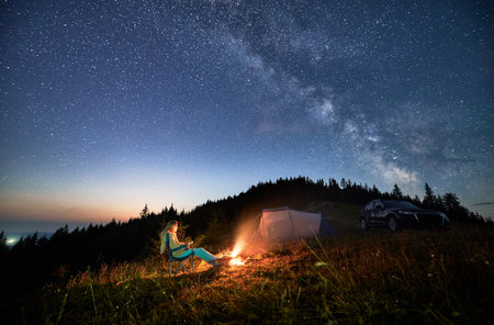 Night camping in the mountains under starry sky full of stars. Travelling woman admiring starry night in mountains. Young woman sitting next to camp fire, tourist tent and off-road car.の写真素材