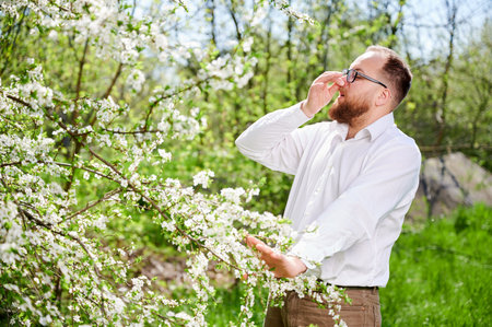 Man allergic suffering from seasonal allergy at spring in blossoming garden at springtime. Young man with glasses sneezing, closing nose by hand in front of blooming tree. Spring allergy conceptの写真素材