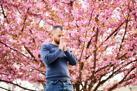 Man allergic suffering from seasonal allergy at spring in blossoming garden at springtime. Man sneezing and blowing nose using nasal handkerchief in front of blooming tree. Spring allergy concept.の写真素材