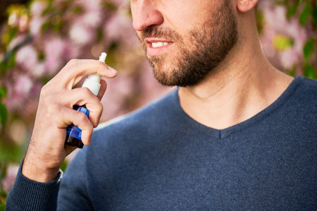 Man allergic using medical nasal drops, suffering from seasonal allergy at spring in blossoming garden. Handsome man treating runny nose in front of blooming tree outdoors. Spring allergy concept.の写真素材