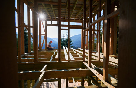 Carpenter constructing wooden frame, two-story house near forest in the morning. Bearded man hammering nails with hammer while dressed in protective helmet and work coveralls at sunrise.の写真素材