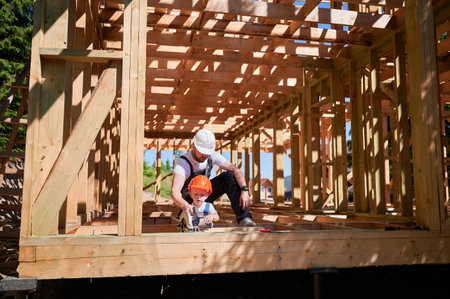 Father with toddler son building wooden frame house. Male builder teaching his son how to cut board using electric saw on construction site, wearing helmets and overalls. Carpentry and family concept.の写真素材