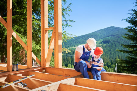 Dad with toddler son constructing wooden frame house. Male builder showing his son the construction plan, wearing helmets and blue overalls on sunny day. Carpentry and family concept.の写真素材