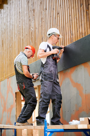 Carpenters constructing wooden framed house. Two men workers cladding facade of house with cement particle boards, fastening them with screwdrivers. Concept of modern eco-friendly construction.の写真素材