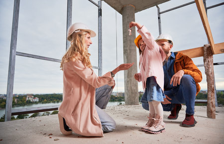 Adorable little girl giving keys from new apartment to mother while standing next to father in building under construction. Happy family spending time together at construction site of new home.の写真素材