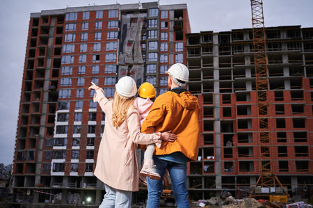Back view of woman pointing at apartment building under constructing while standing next to husband and daughter on the street. Happy family looking at new home at construction site.の写真素材