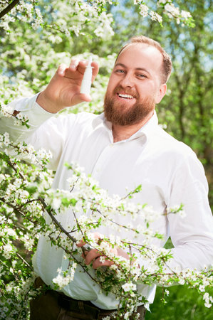 Man allergic using medical nasal drops, suffering from seasonal allergy at spring in blossoming garden. Portrait of smiling man showing medicament near blooming tree outdoors. Spring allergy concept.の写真素材