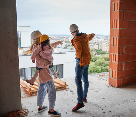 Back view of parents with daughter standing inside apartment building under construction. Man and woman with child wearing building helmets while enjoying city view from future home.の写真素材