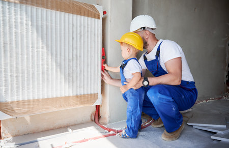 Father and little son using spirit level tool while installing heating radiator in apartment. Male plumber and child measuring pipes level with building instrument.の写真素材