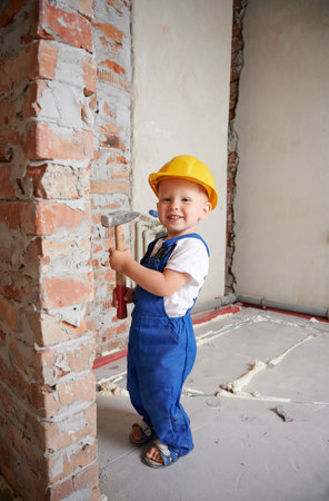 Full length of happy child construction worker holding hammer tool and looking at camera while standing by brick wall at home during renovation. Smiling kid wearing safety helmet and work overalls.の写真素材