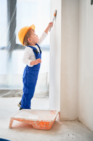 Full length of child construction worker painting wall with paint brush in living room. Kid in safety helmet and work overalls using paint brush while working on apartment renovation.の写真素材