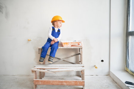 Cute kid construction worker sitting on wooden table against white wall in apartment under renovation. Cheerful little boy wearing safety helmet and work overalls while playing at home.の写真素材
