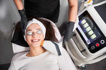 Top view of cheerful woman in safety goggles receiving laser facial treatment in cosmetology clinic. Beautician using diode laser scanner device while performing resurfacing skincare procedure.の写真素材