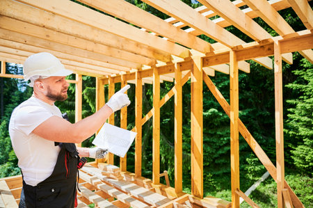 Surveyor inspecting construction plans of wooden two-story building being erected. Bearded man in jumpsuit and helmet pointing on the wall. Concept of contemporary ecological construction methods.の写真素材