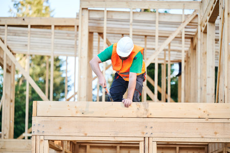 Carpenter constructing two-story wooden frame house. Bearded man hammering nails into structure while wearing protective helmet and construction vest. Concept of modern ecological construction.の写真素材