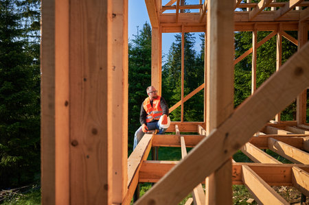 Carpenter constructing wooden frame two-story house near the forest. Bearded man holding hammer, dressed in protective helmet and orange safety vest. Concept of ecological modern construction.の写真素材
