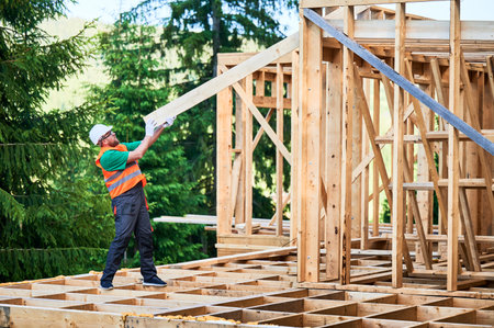 Carpenter constructing wooden-framed house near the woods. Man holding large truss, wearing work clothes and helmet. Concept of modern and eco-friendly building.の写真素材