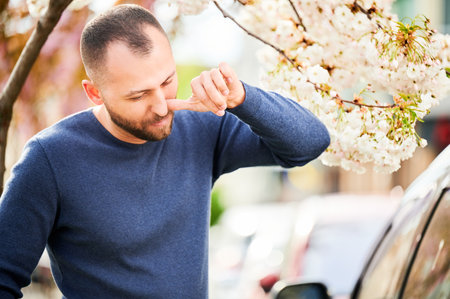 Man allergic suffering from seasonal allergy at spring in blossoming garden at springtime. Bearded young man sneezing and blowing runny nose in front of blooming tree. Spring allergy concept.の写真素材