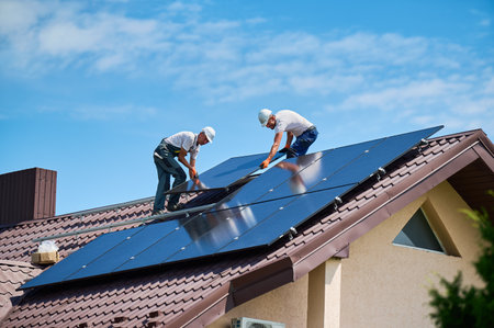 Workers building solar panel system on rooftop of house. Two men installers in helmets installing photovoltaic solar module outdoors. Alternative, green and renewable energy generation concept.の写真素材