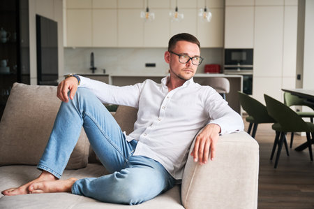 Charming man relaxing on couch in his apartment. Handsome young male thoughtfully looking away. Man in casual clothes lying on sofa on background of modern kitchen.の写真素材