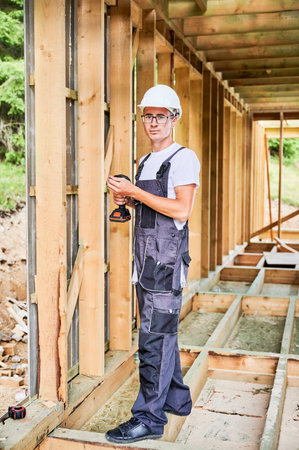 Carpenter constructing wooden framed house. Man worker in glasses working with screwdriver, wearing work overalls and helmet. Concept of modern eco-friendly construction.の写真素材