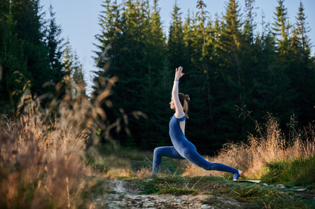 Young woman doing warrior one pose. Sporty female practicing new yoga pose. Meditation at sunrise in forest.の写真素材