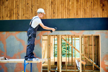 Carpenter constructing wooden framed house. Man worker in glasses cladding facade of house with cement particle boards, fastening them with screwdrivers. Concept of modern eco-friendly construction.の写真素材