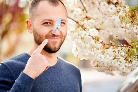 Man allergic suffering from seasonal allergy at spring. Bearded man with clothespin clipped to his nose - symbolic gesture of his inability to breathe due to nasal congestion near blooming tree.の写真素材
