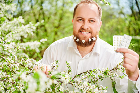 Man allergic suffering from seasonal allergy at spring. Handsome man with small white flowers dotted in beard, holding pack of pills, posing in blossoming garden. Antihistamine medication conceptの写真素材