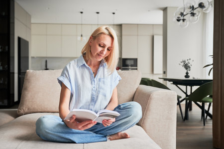 Woman sitting comfortably on couch, deeply engaged in reading book. Cozy living space with modern aesthetic on background. Person enjoying quiet moment of solitude, immersed in reading.の写真素材